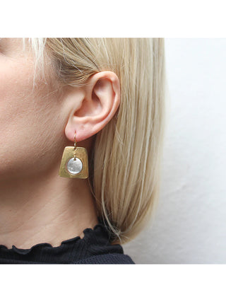 A brass cutout tapered rectangular earring with a silver-toned disc hanging in the center, on a model with blond hair. 