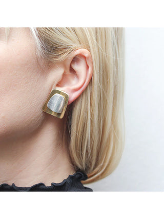 An earring of lightly dished brass tapered rectangle layered with a silver-toned, domed tapered rectangle, on a blond-haired model's ear. 