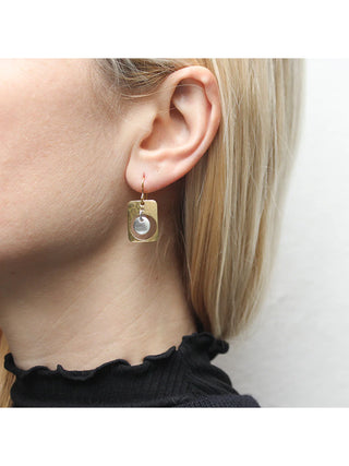 Earring with a brass cutout rounded rectangle with a silver-toned disc hanging in the center, on a blond-haired model's ear.