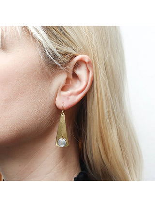 A brass cutout long triangle earring with a silver disc hanging in the center, on a blond-haired model's ear. 