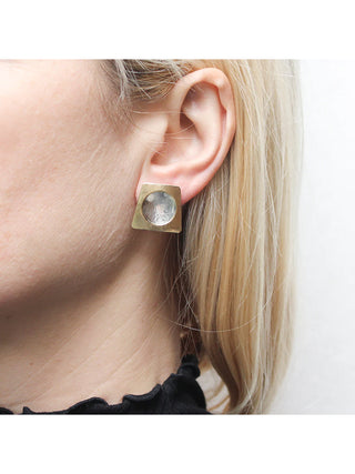 A model's ear with a a brass tapered rectangular earring with a cutout in the middle layered over a dished silver-toned disc.