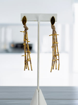 The earrings, made of a gold-plated leaf-shaped piece with four gold colored cords hanging from it, holding bronze gold-plated ingots, displayed on a blurred background.