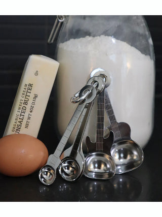 A set up stainless steel guitar-shaped spoons leaning against a jar of flower with a stick of butter and egg adjacent.