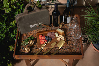 A wooden table with a loaded charcuterie board, the travel cheese knife set and its case, and two wine glasses next to bottles of wine.