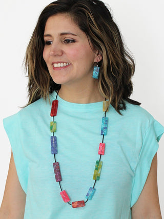 A model in a light blue top wearing A necklace with rectangular resin beads in rainbow colors from red to blue to green to purple, on a cord.