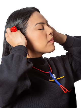 A model in a black top with her eyes closed, wearing a necklace with a red chain and a little man pendant comprised of blue, yellow and red, with a clear head. She's also wearing a red squiggly ring.