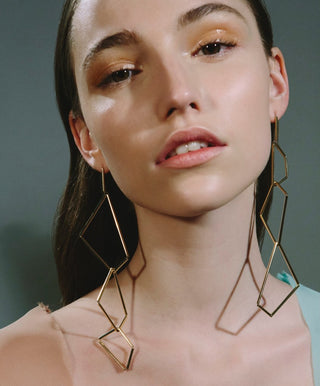 A pair of long gold earrings with an origami-like shape, on a model looking directly ahead.