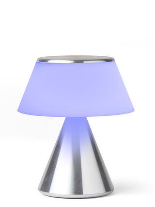 A lamp with a triangular aluminum base and an illuminated  lilac shade.