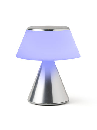 A lamp with a triangular aluminum base and an illuminated lilac shade.