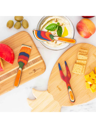 Three wooden cheese tools in vibrant colors laying on the table with a charcuterie spread.