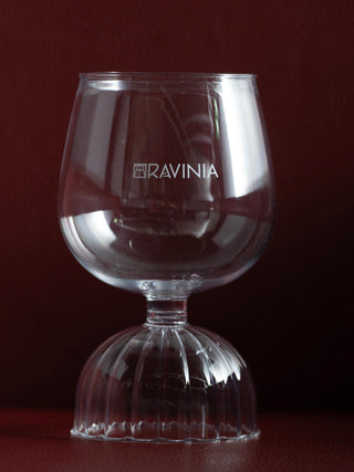 The clear Milan red wine glass with the Ravinia name and logo displayed upon a dark red background.