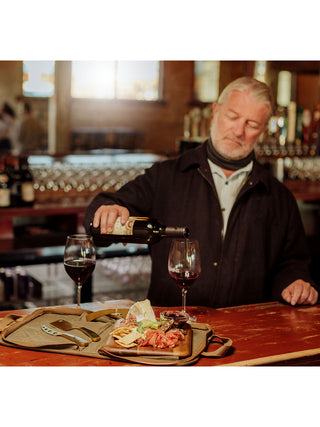 A gray haired man with a beard pouring wine into a glass, with a cheese board full of appetizers and cheese knives adjacent in the foreground.