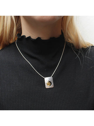 A necklace with a silver-toned cutout and rounded rectangle with a brass hanging disc in the bottom center, on a silver-toned snake chain.  A model with a black top is wering it.