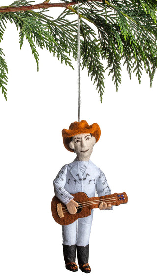 A holiday ornament decicting Hank Williams in a cowboy hat with a guitar, hanging from a Christmas tree branch.