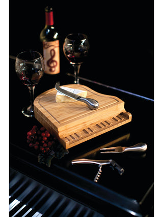 A tiny wooden piano cheese board on top of a real piano, with two glasses of wine, cheese, cheese knives, and a corkscrew.