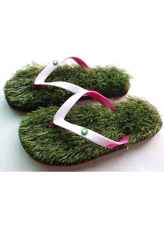 A pair of flip flops that appear to be made of grass, with pinks and white straps, standing leaning against a wood board next to a swimming pool.