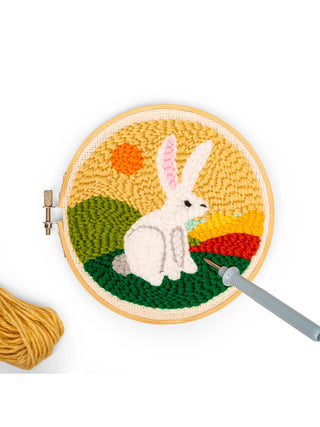 A finished punch needle kit in a wooden embroidery hoop depicting a white hare, with a punch needle and some yarn next to it.