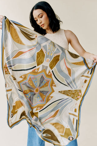 A woman holding out a square scarf that has patterns in amber, blue, and off-white.
