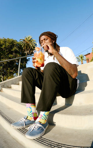 A young man witting on steps with a drink, wearing A flat pair of socks with smiley faces and colorful rainbow checker designs.