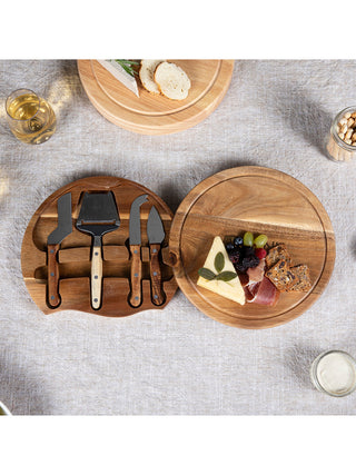 The acacia wood cheese board with some cheese and crackers, fully open to reveal the four cheese tools, all on a gray tablecloth with various food items around it.