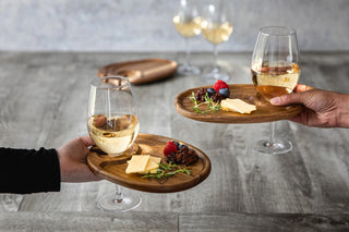 Two hands holding wooden appetizer plates with food, and stemmed wine glasses held in the built-in space, all on a gray background.