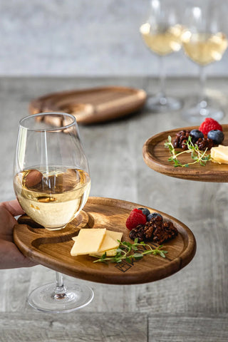 A hand holding a wooden appetizer plate with food, and a stemmed wine glass held in the built-in space, all on a gray background.