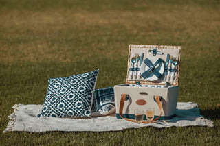A blanket laying on a field, with pillows and a picnic basket for 2.