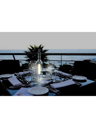 A lit bottlelight sitting on a table for four at dusk, in front of a body of water.