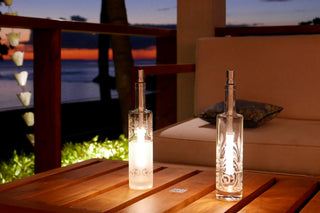 An outside dining area by the beach, with two bottlelight lamps in decorated glass bottles sitting on a wooden table.