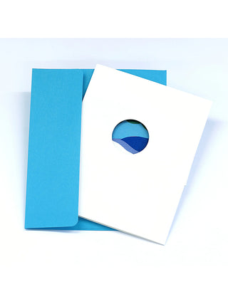 A white greeting card with a round cutout in the center, in front of a blue envelope.