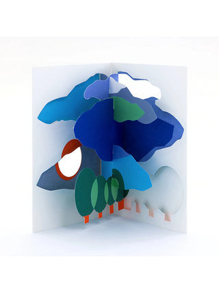 An open white pop-up card with blue clouds and green trees.