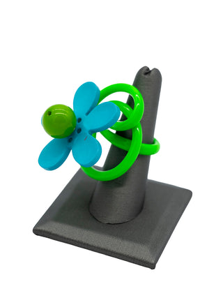 A green squiggle ring with a turquoise and green flower, displayed on a gray ring holder.
