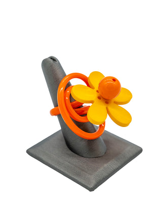A orange squiggle ring with a yellow and orange flower, displayed on a gray ring holder.