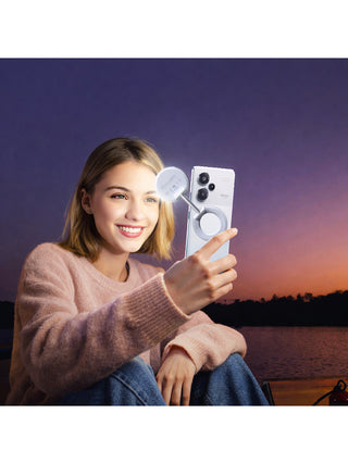 A person taking a selfie at dusk, holding a phone with the MagSafe compatible GloLens Pro light atttached.