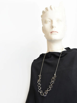 A mannequin wearing a black shirt and a satin gold long necklace with a section of linked cube pieces in the center.