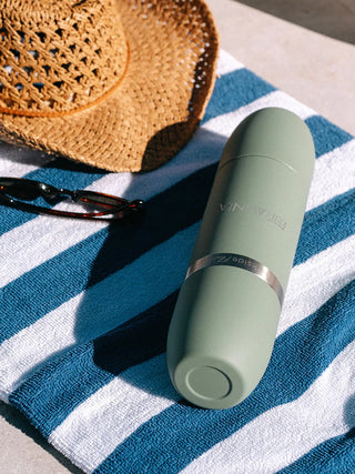 A green wine flask laying on a blue and white blanket, next to a straw hat and sunglasses.