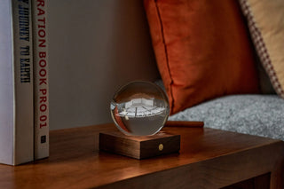 A crystal globe etched with the Pavilion sitting on a lit wooden base, on a wooden bedside table.