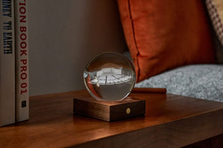 A crystal globe etched with the Pavilion sitting on a lit wooden base, on a wooden bedside table.