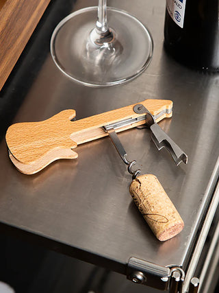 A guitar-shaped corkscrew sitting on a metal counter, next to a wine glass and a bottle of wine.