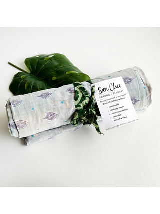 A rolled sari blanket with a green fabric tie and a Sari Chic Sarong + Blanket tag, on a white table in front of a green leaf.