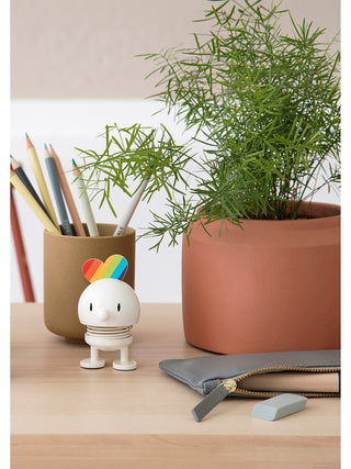 A smiling white figure with a spring in its middle and a rainbow behind its head, adjacent to a potted plant and in front of a container of pencils.