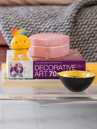 A yellow plastic figure with a spring in its middle perched on a Decorative Art book.