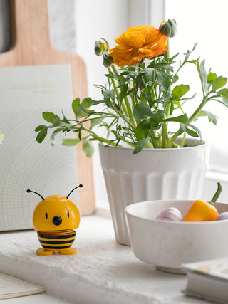 A bee-like figure with a spring in its middle, posed next to a flower pot on a window sill.