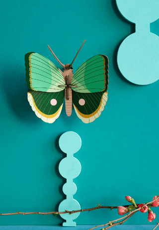 A cardboard butterfly with a brown body and two shades of green on its wings., mounted on a teal-colored wall.