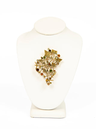 A gold brooch with leaves, sheet music and multicolored crystals on a form.