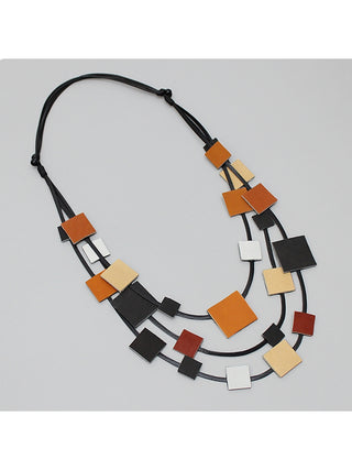 An angled view of a necklace features multiple strands of black cord with square leather pieces in shades of brown and black, strung throughout. 