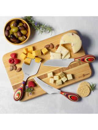 A cheese boars covered with cheese and meat, and three knives resting on it, a bowl of olives adjacent.