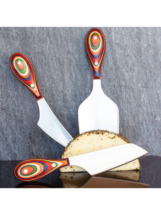 Three cheese knives with color handles poised next to a block of cheese.