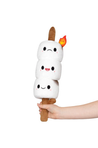 A hand holding a stick with three plush marshmallows with faces, on a stick, the top one frowning at a flame begins to burn him.