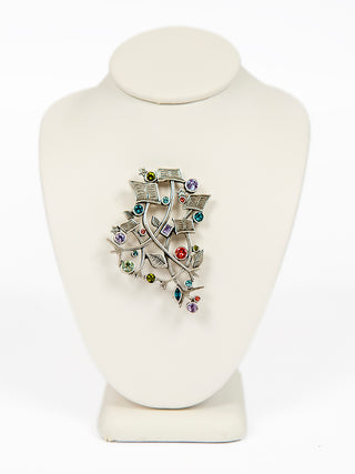 A silver brooch with leaves, sheet music and multicolored crystals on a form.
